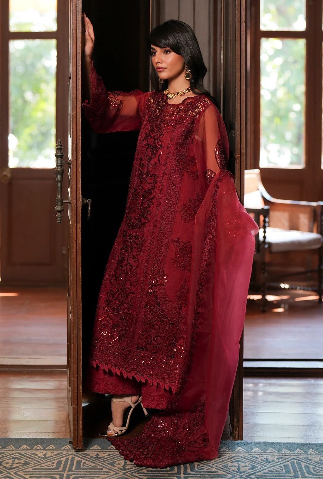 A woman models a deep maroon red embroidered traditional outfit with sheer sleeves and a matching dupatta, standing in a doorway of a wooden house.