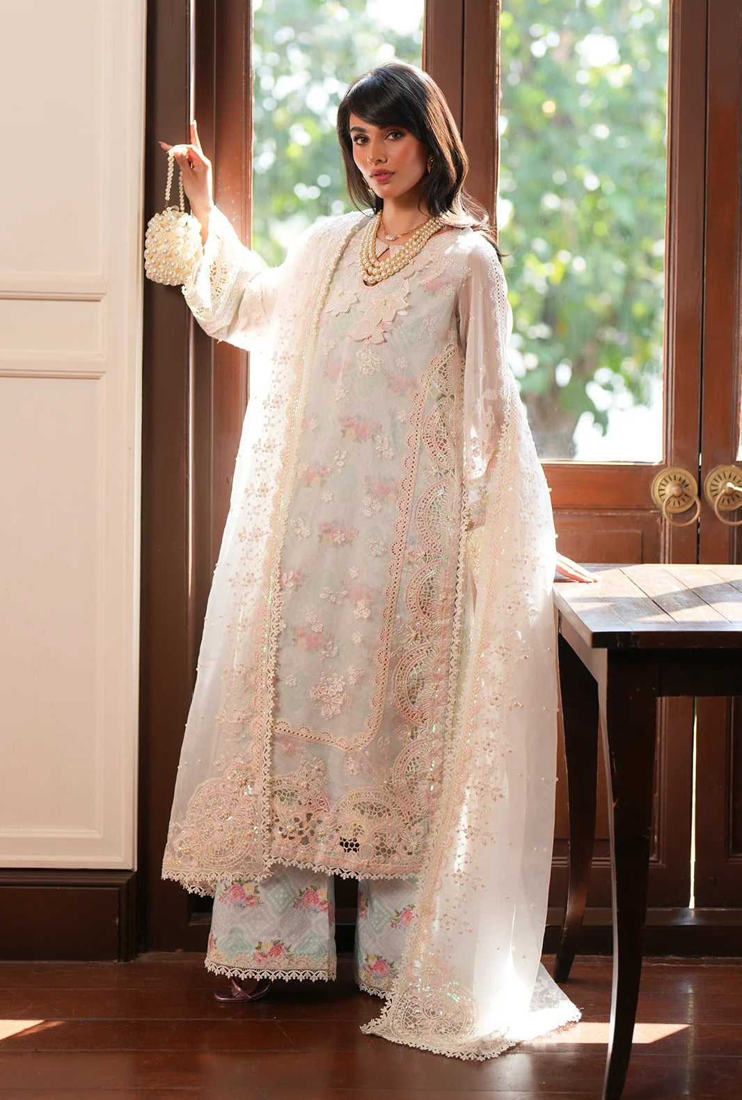 A woman models a traditional white and light blue embroidered South Asian outfit with a matching sheer dupatta, standing in a sunlit room with wooden doors and a table.