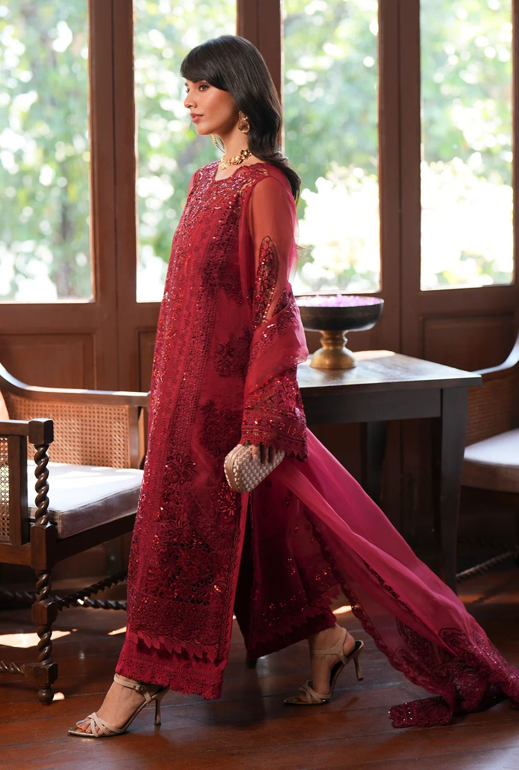 A woman models a deep red embroidered traditional outfit with a long sheer overlay and matching skirt in an indoor setting with wooden furniture and large windows.