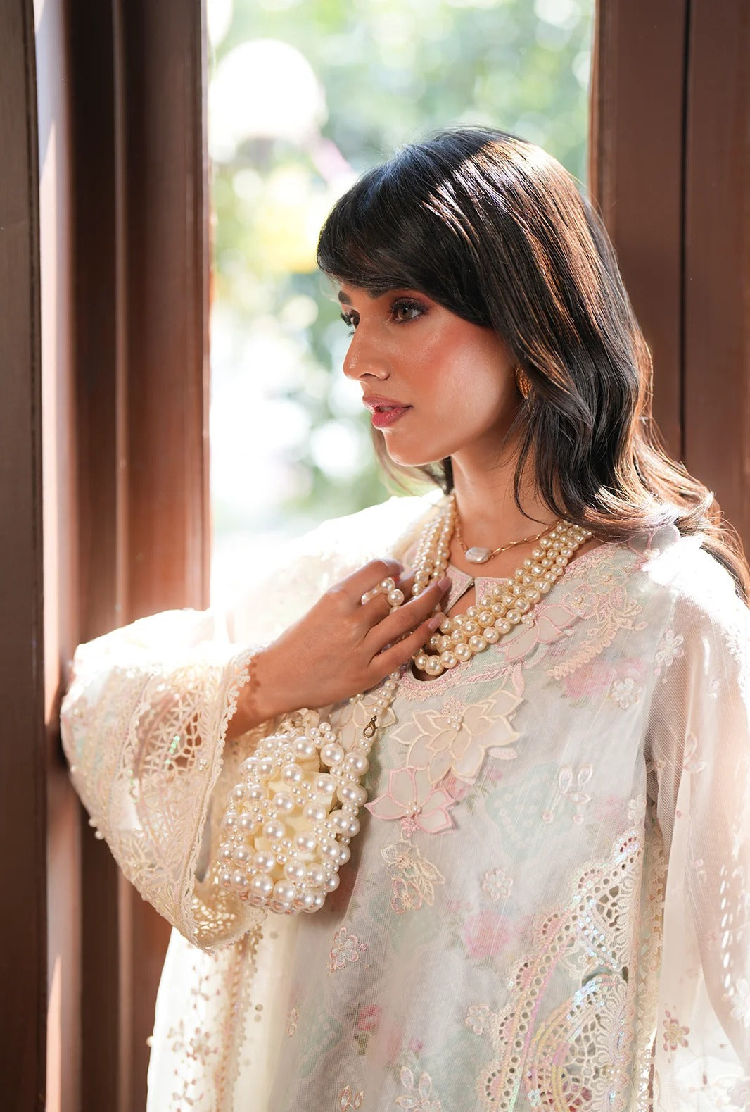A woman wearing a cream-colored embroidered traditional outfit with pearl necklaces and earrings, standing near a window with warm brown wooden frames.