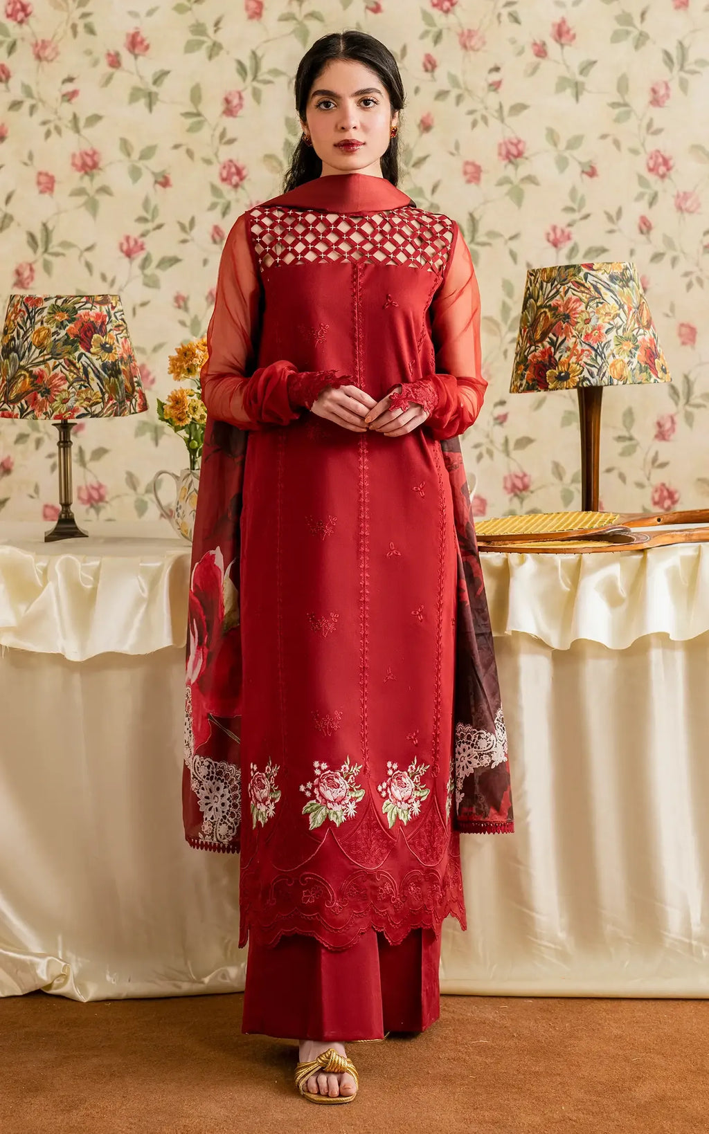 Woman wearing a red embroidered dress in a decorated room with floral wallpaper and lamps.