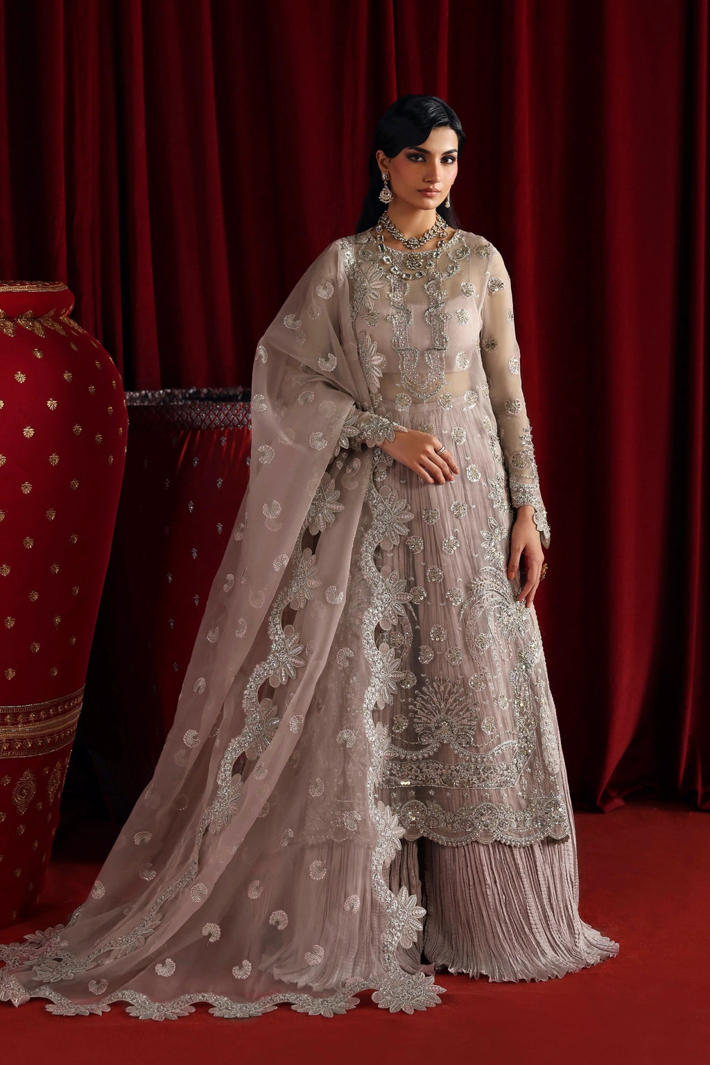 A woman models a traditional Indian salwar kameez outfit in a dusty rose/taupe color with intricate silver embroidery, paired with a matching sheer dupatta, set against a dark red curtain backdrop.