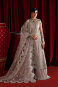 A woman models a traditional Indian salwar kameez outfit in a dusty rose/taupe color with intricate silver embroidery, paired with a matching sheer dupatta, set against a dark red curtain backdrop.