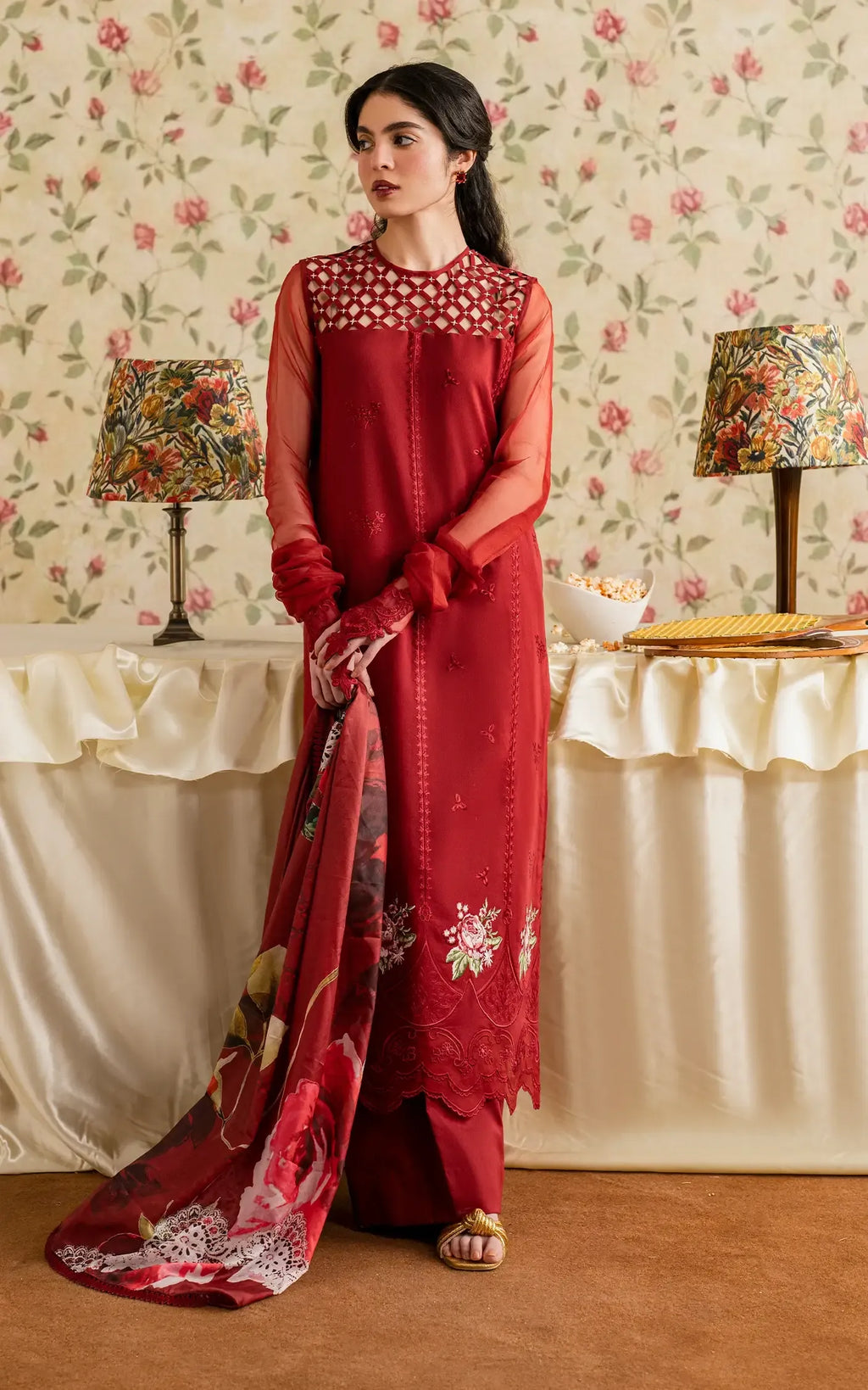 Woman in a red traditional outfit standing in a room with floral wallpaper and lamps.