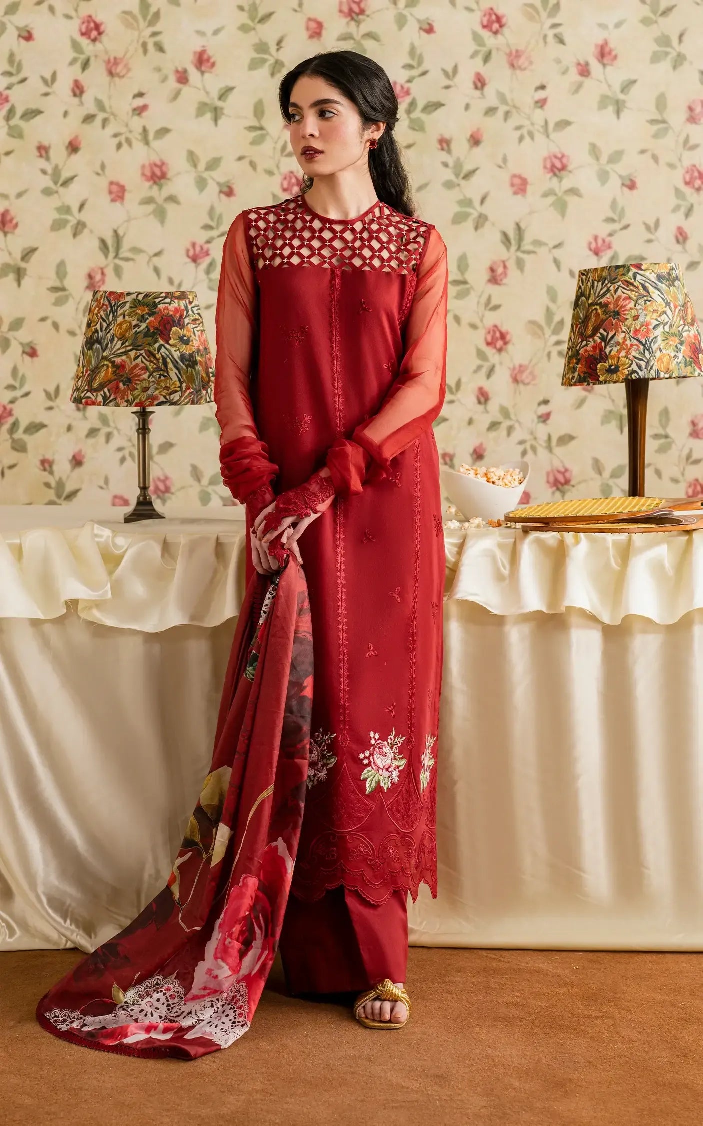 Woman in a red traditional outfit standing in a room with floral wallpaper and lamps.