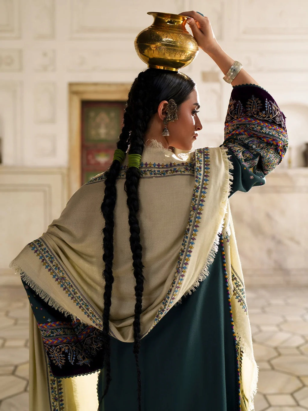 A woman in traditional South Asian attire, viewed from behind, balancing a brass pot on her head. She wears a dark teal dress, a beige shawl with colorful embroidery, and a dark embroidered jacket.