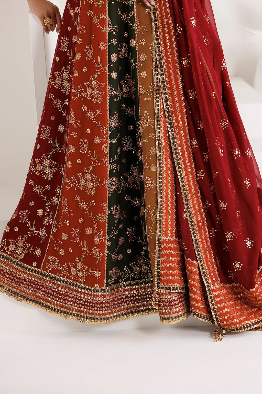 A close-up of a traditional Indian ethnic garment, likely a lehenga or skirt, featuring three vertical panels in rust red, black, and brown with intricate gold floral embroidery and a heavily embellished border. A matching red dupatta is draped over the side, decorated with small gold mirror work and floral patterns.