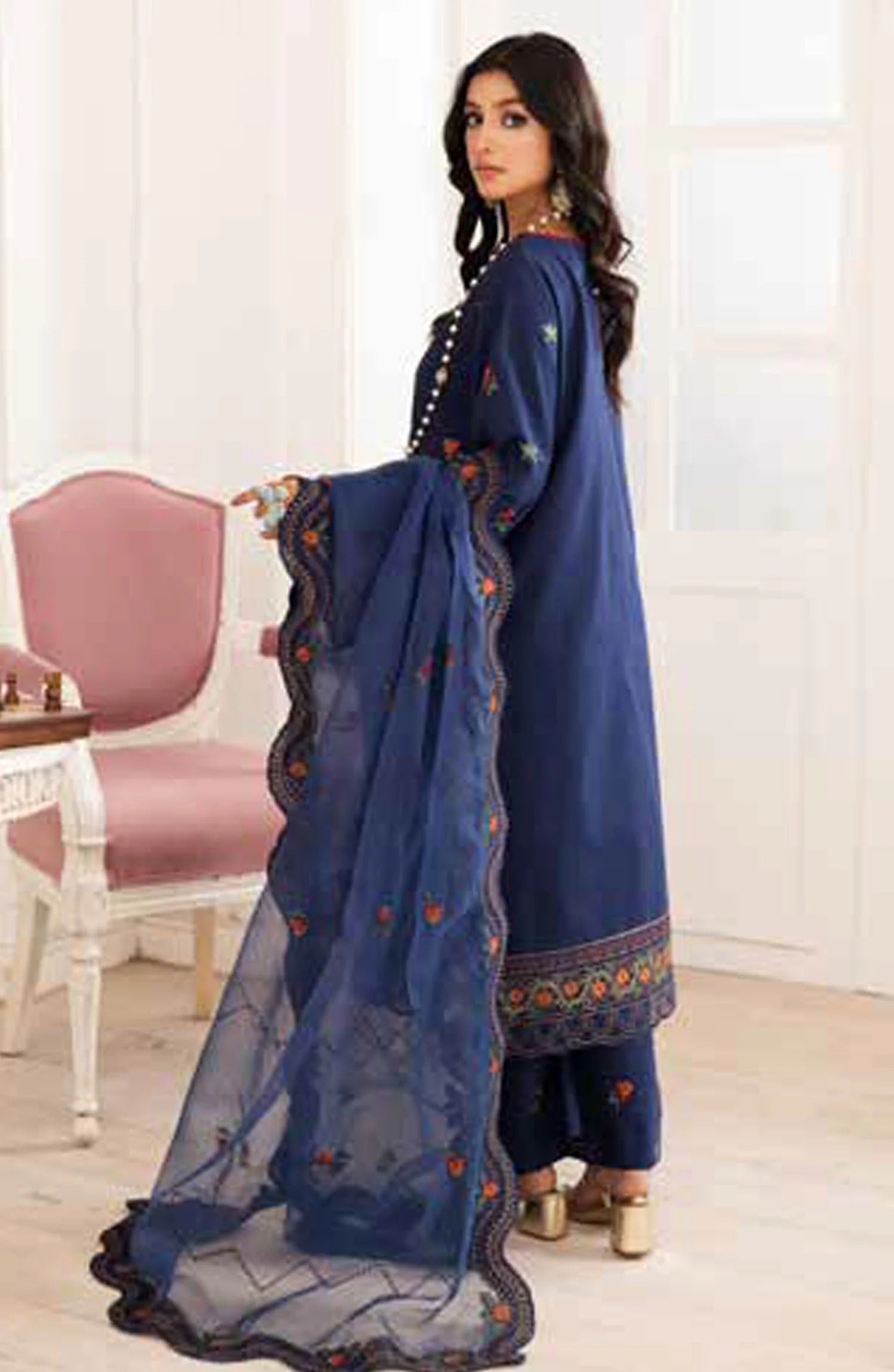 A woman models a traditional navy blue ethnic suit set featuring a long kurta, matching pants, and a sheer dupatta with floral embroidery and scalloped borders, set against an indoor background with a pink chair.