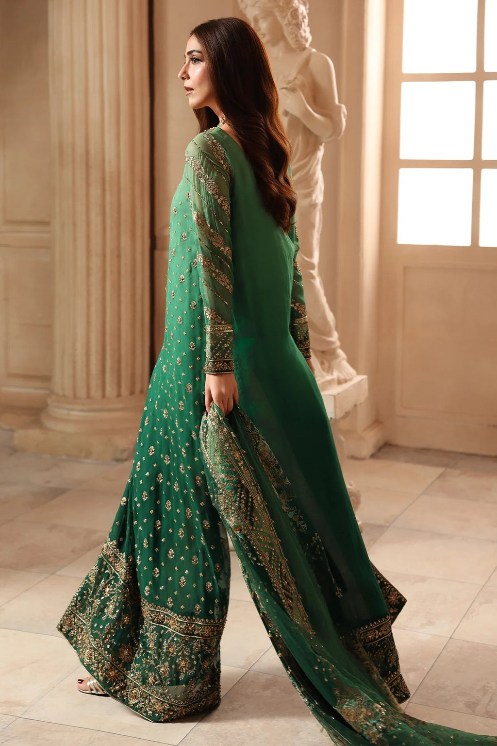 A woman wearing a green traditional outfit with gold embroidery, standing in a classical interior with columns and a statue.