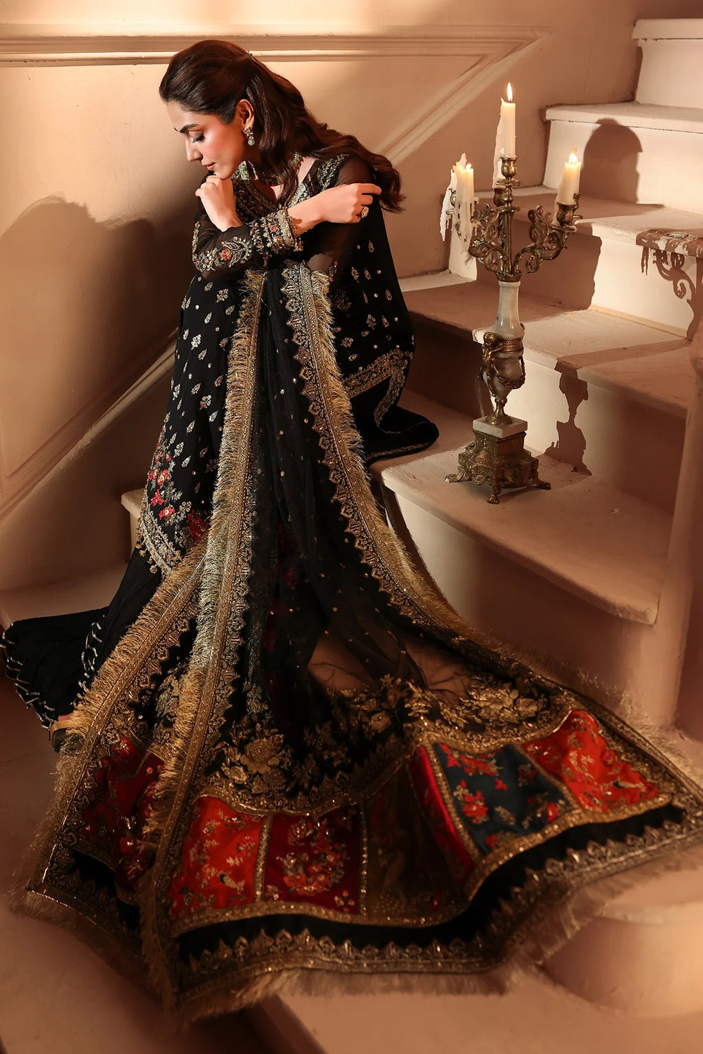 A woman wearing a black embroidered traditional outfit with a heavily embellished dupatta and red patterned lower panel, posed on an indoor staircase beside lit candles.