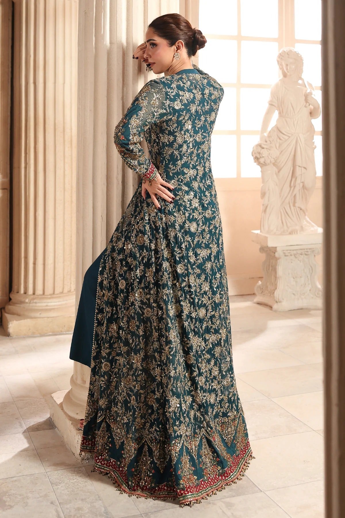 A woman modeling a long teal blue and beige floral printed anarkali gown in an elegant indoor setting with classical columns and a white statue in the background.