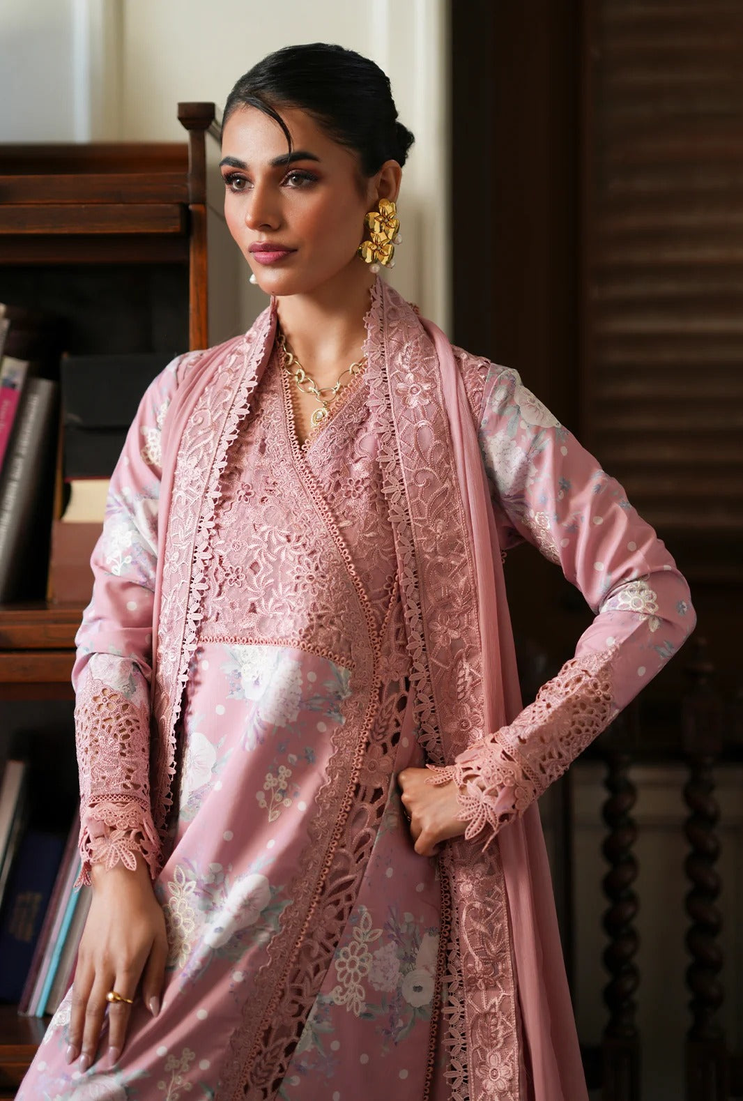 A woman models a traditional pink embroidered outfit with matching scarf in an indoor setting with a wooden bookshelf in the background.
