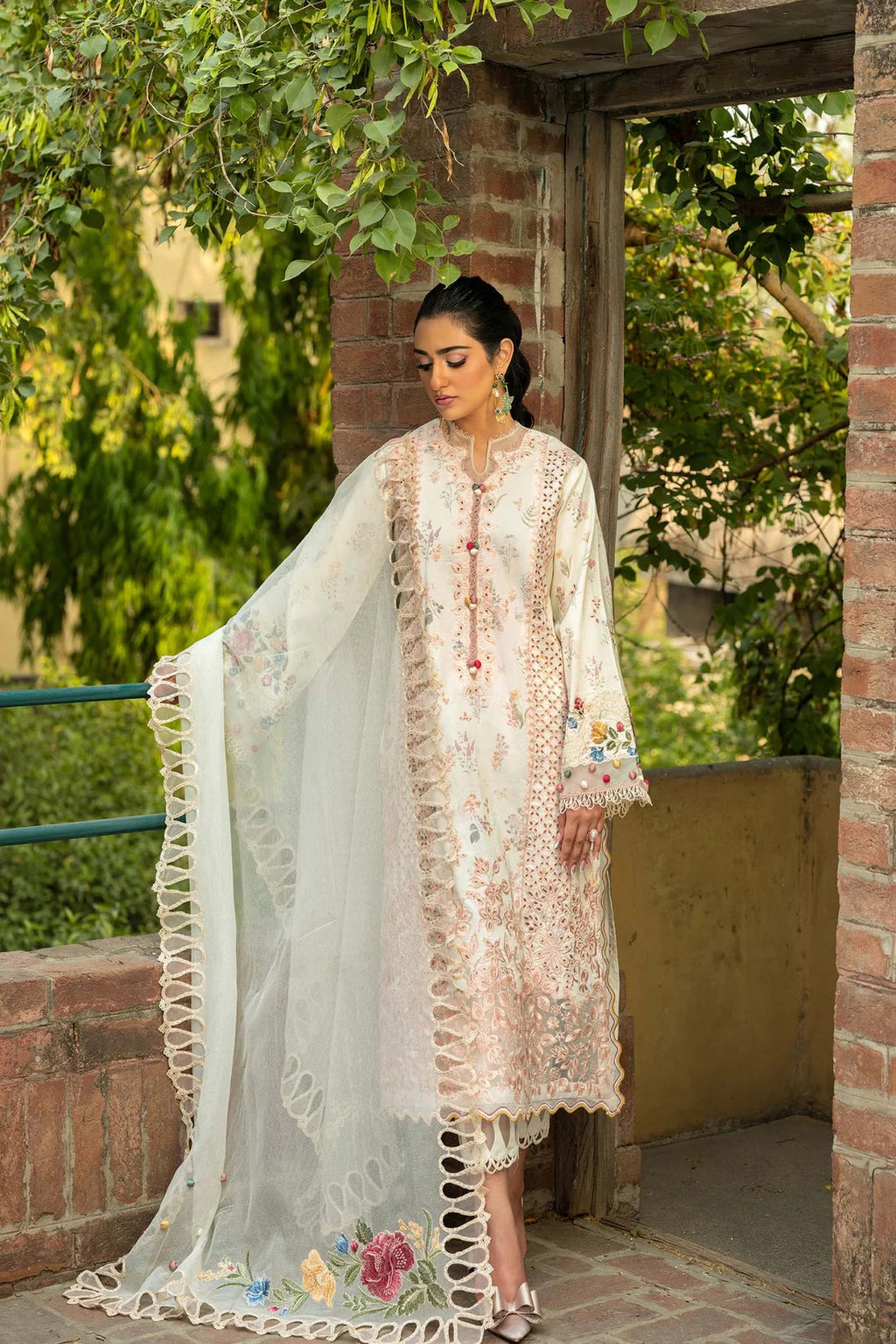 A woman models a traditional three-piece salwar kameez suit in a rustic outdoor setting with brick pillars and green foliage. The suit is light beige with intricate floral embroidery and features a matching sheer dupatta with scalloped edges and floral prints.