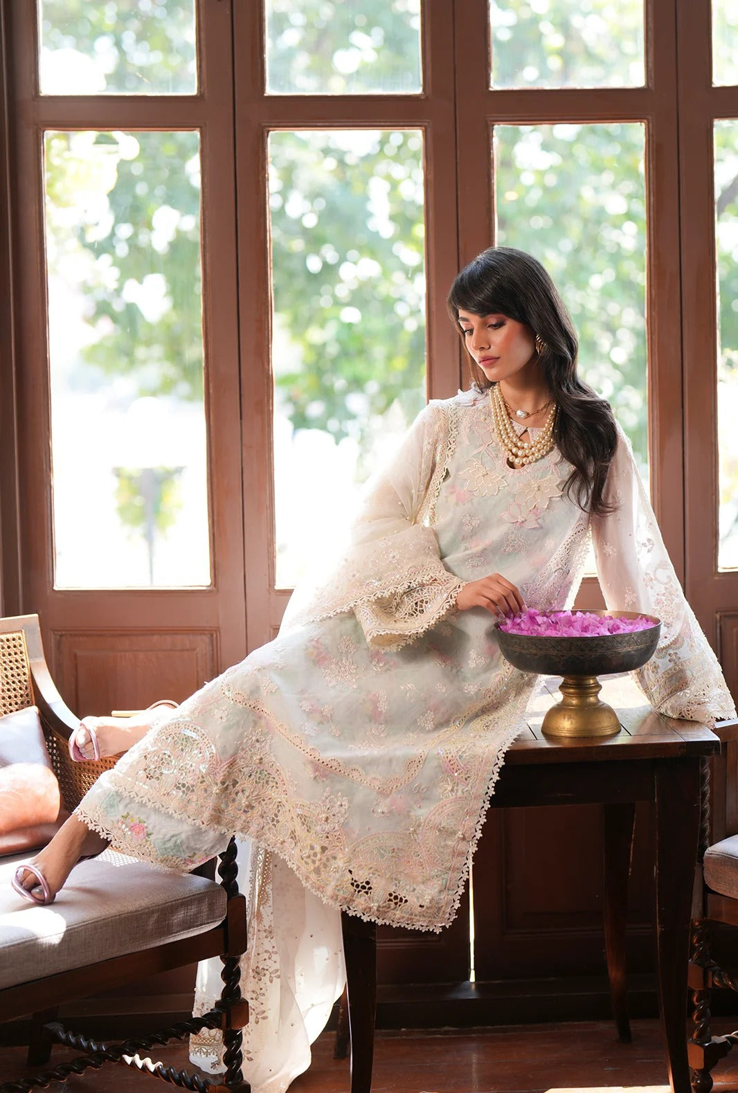 A woman wearing a traditional white embroidered kurta with a matching dupatta and gold jewelry sits on a wooden bench in a sunlit room with large windows.