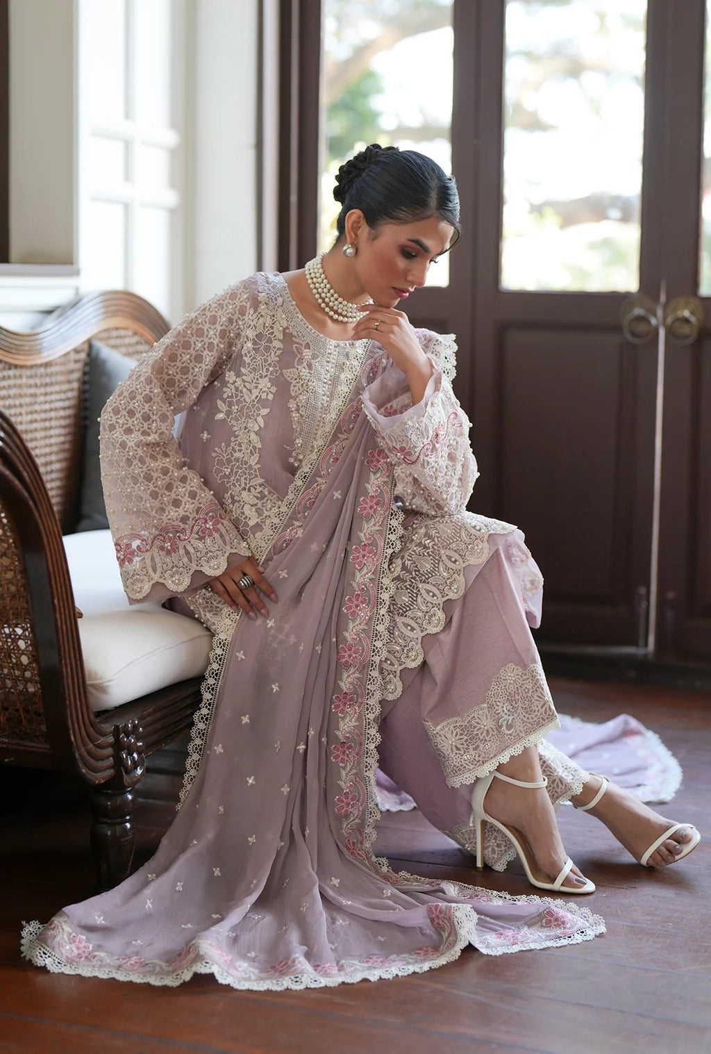 A woman seated indoors wearing an elegant lavender traditional outfit with intricate white floral embroidery and lace detailing, paired with white heeled sandals and jewelry.