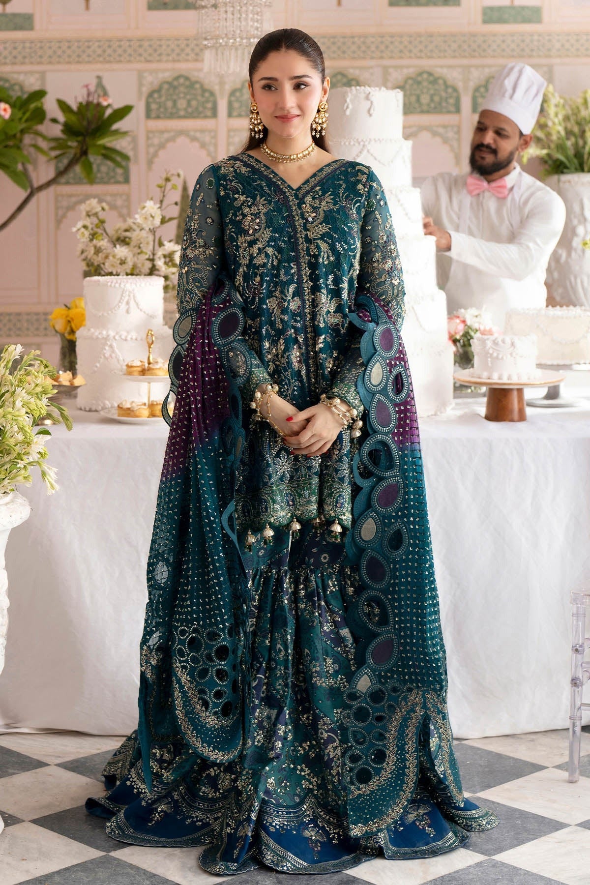 A woman wearing an ornate teal and purple embroidered traditional gown with gold detailing, standing in an elegant indoor setting with decorated cakes and a chef in the background.