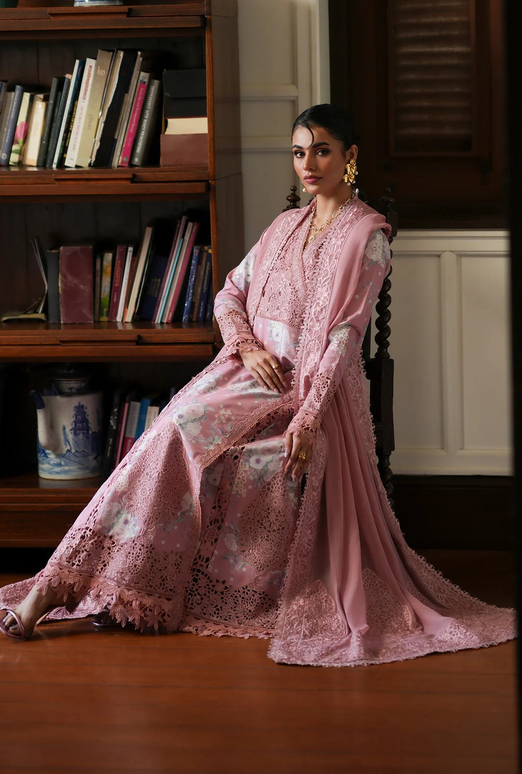 A woman wearing a long pink embroidered traditional outfit with sheer sleeves and a flowing skirt, seated in a room with wooden bookshelves and a wood floor.