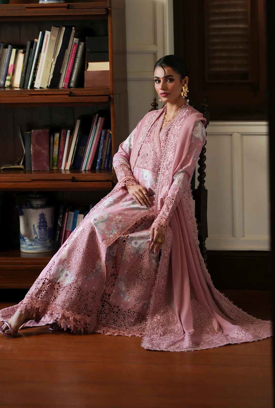 A woman wearing a long pink embroidered traditional outfit with sheer sleeves and a flowing skirt, seated in a room with wooden bookshelves and a wood floor.