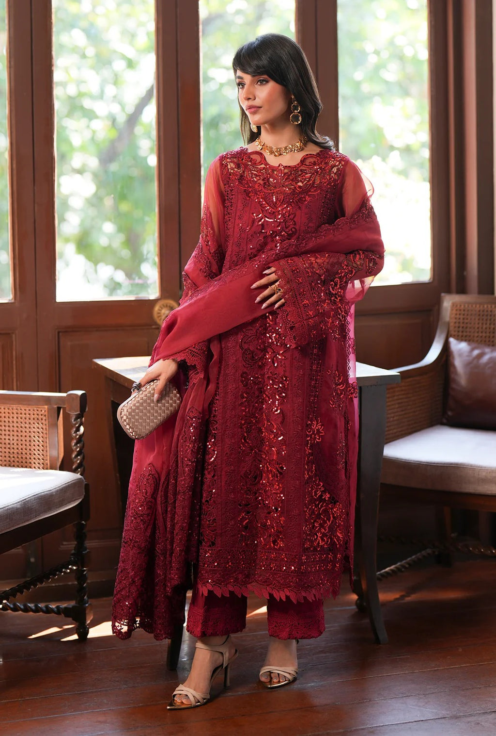 A woman models a deep red embroidered traditional outfit with matching dupatta and trousers, standing in a furnished room with wooden floors and large windows.