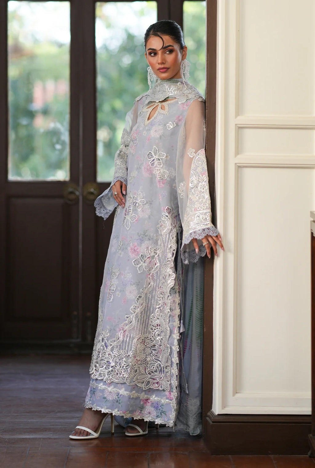A woman models a traditional light blue-gray embroidered dress with floral lace detailing, a matching sheer dupatta, and a gray skirt, standing in an indoor setting with a dark wooden door and cream-colored wall paneling.