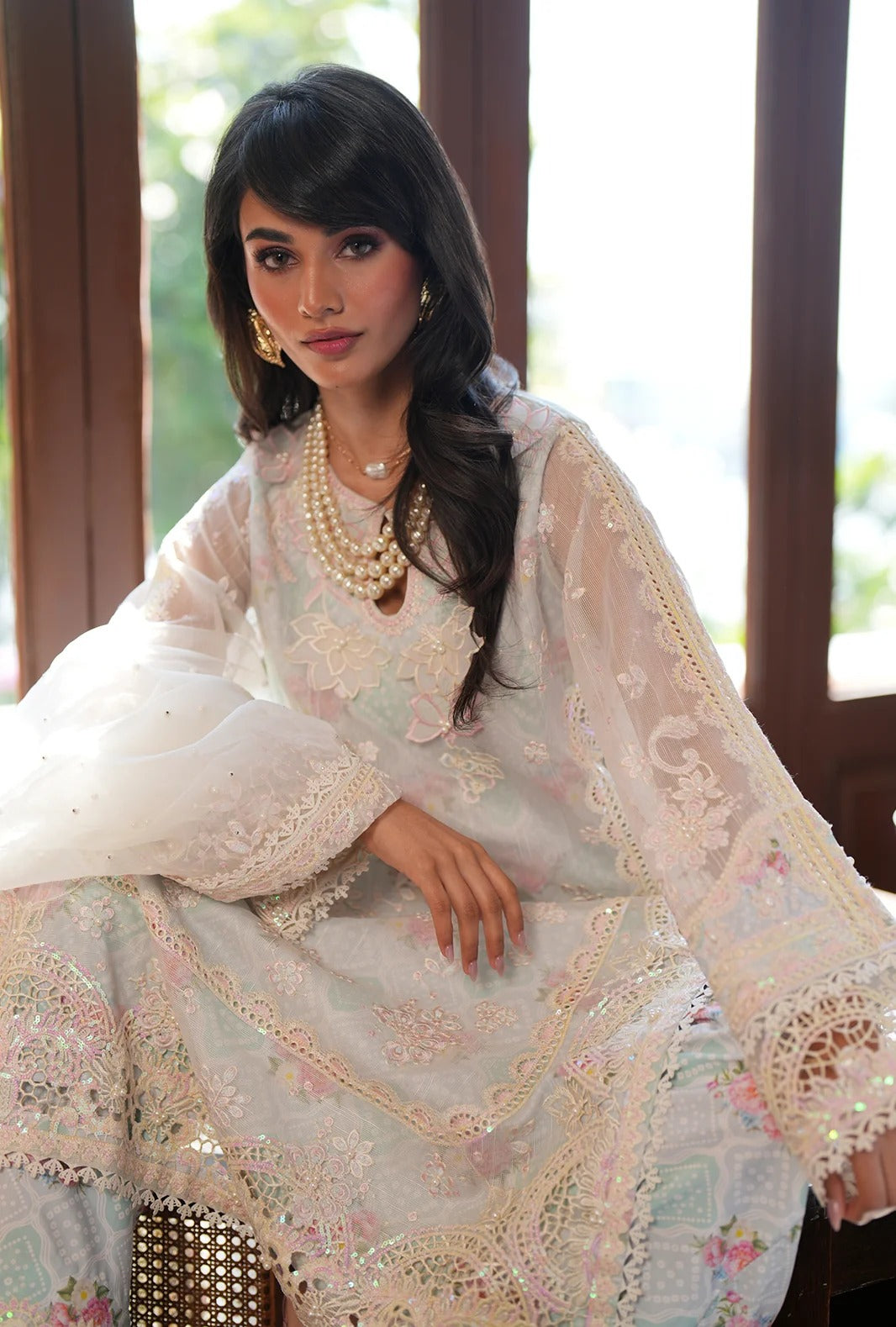 A woman wearing a traditional embroidered white and pale pink ethnic outfit with lace sleeves and a matching dupatta, seated indoors near large windows.