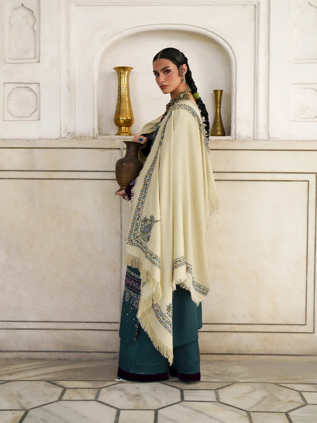 A woman models a traditional South Asian outfit featuring a cream-colored embroidered shawl draped over a dark teal kurta and wide-leg trousers, standing in front of a beige stone wall with decorative arches and gold vases.