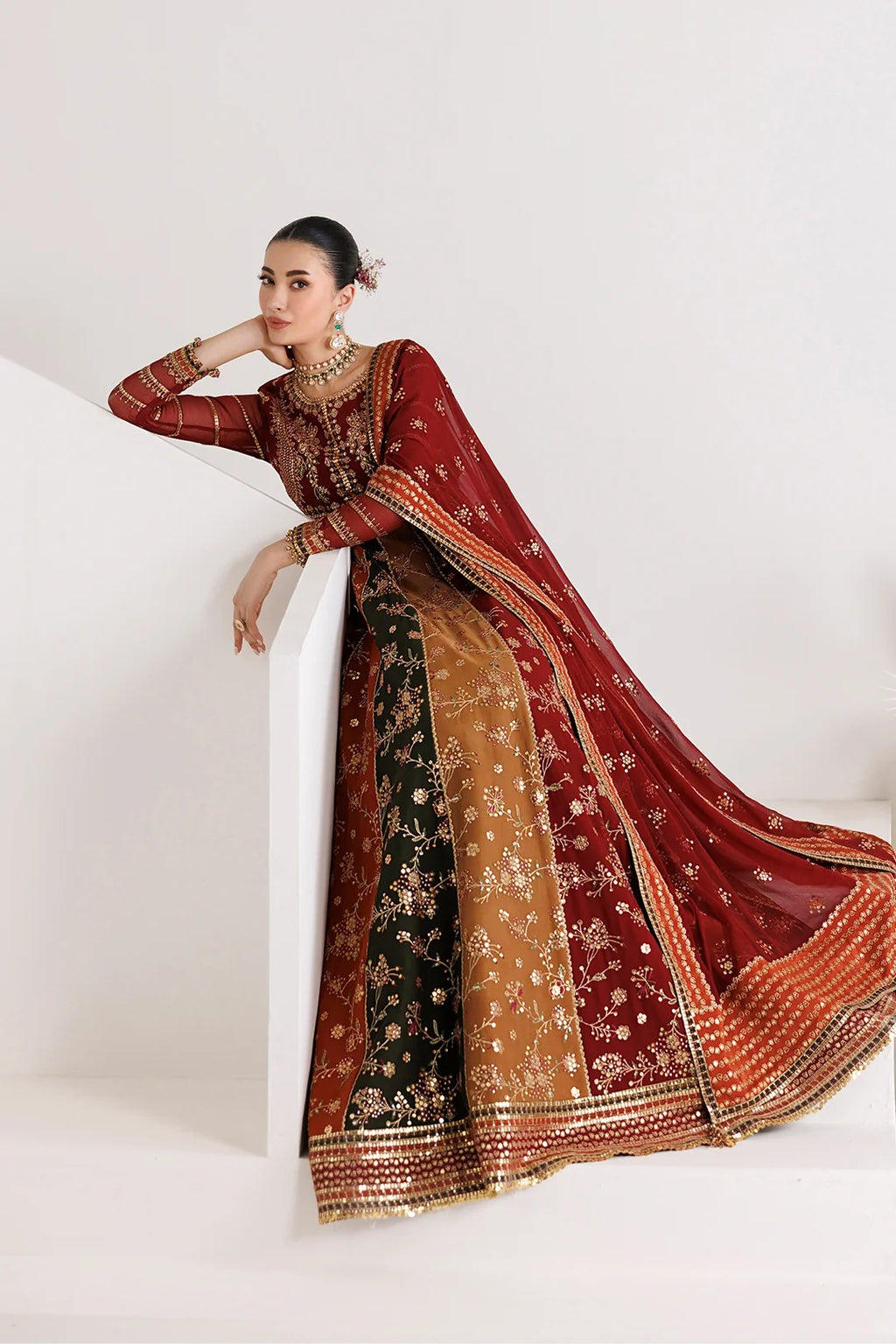 A woman models an ornate traditional Indian lehenga choli set with a deep red dupatta, featuring gold embroidery, a brown embroidered skirt, and a black embroidered panel, posed on white architectural steps against a white background.