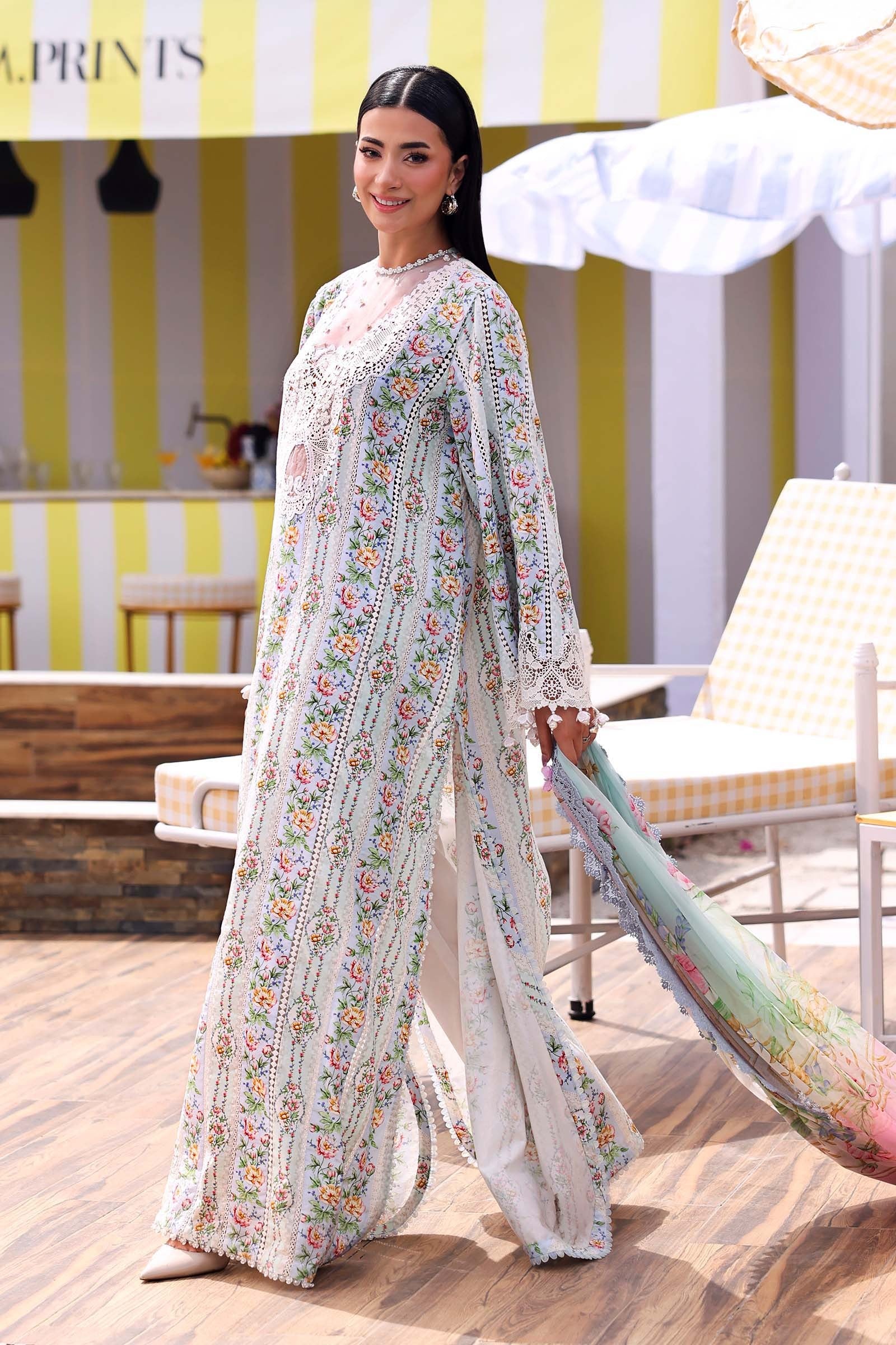 Woman in a floral outfit holding a scarf in an outdoor setting with striped walls.