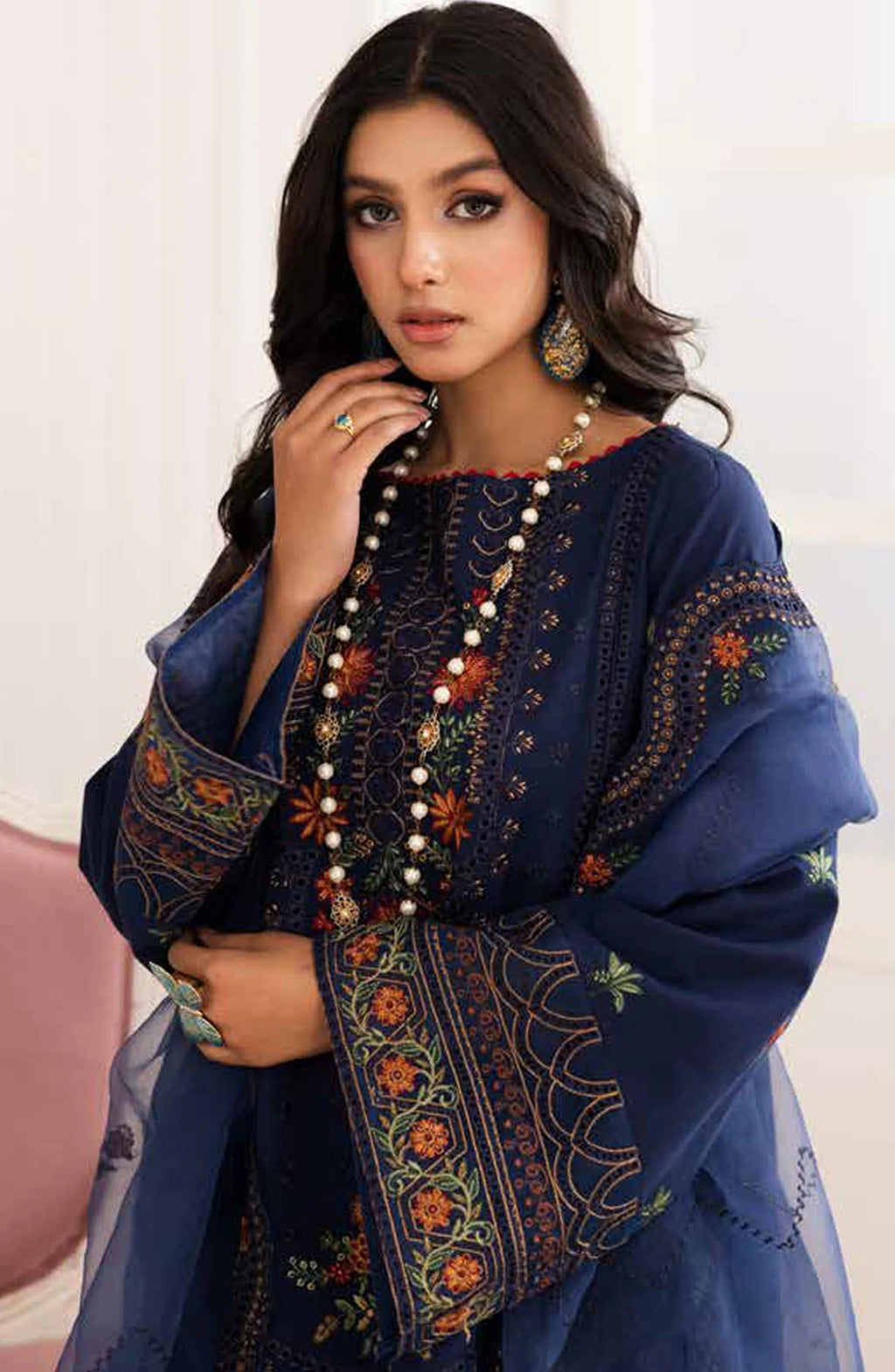 A woman wearing a navy blue embroidered traditional outfit with sheer sleeves, ornate multicolor floral embroidery, and a long beaded necklace, posed against a light indoor background.