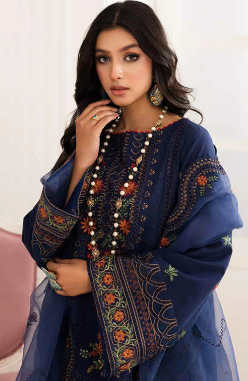 A woman wearing a navy blue embroidered traditional outfit with sheer sleeves, ornate multicolor floral embroidery, and a long beaded necklace, posed against a light indoor background.