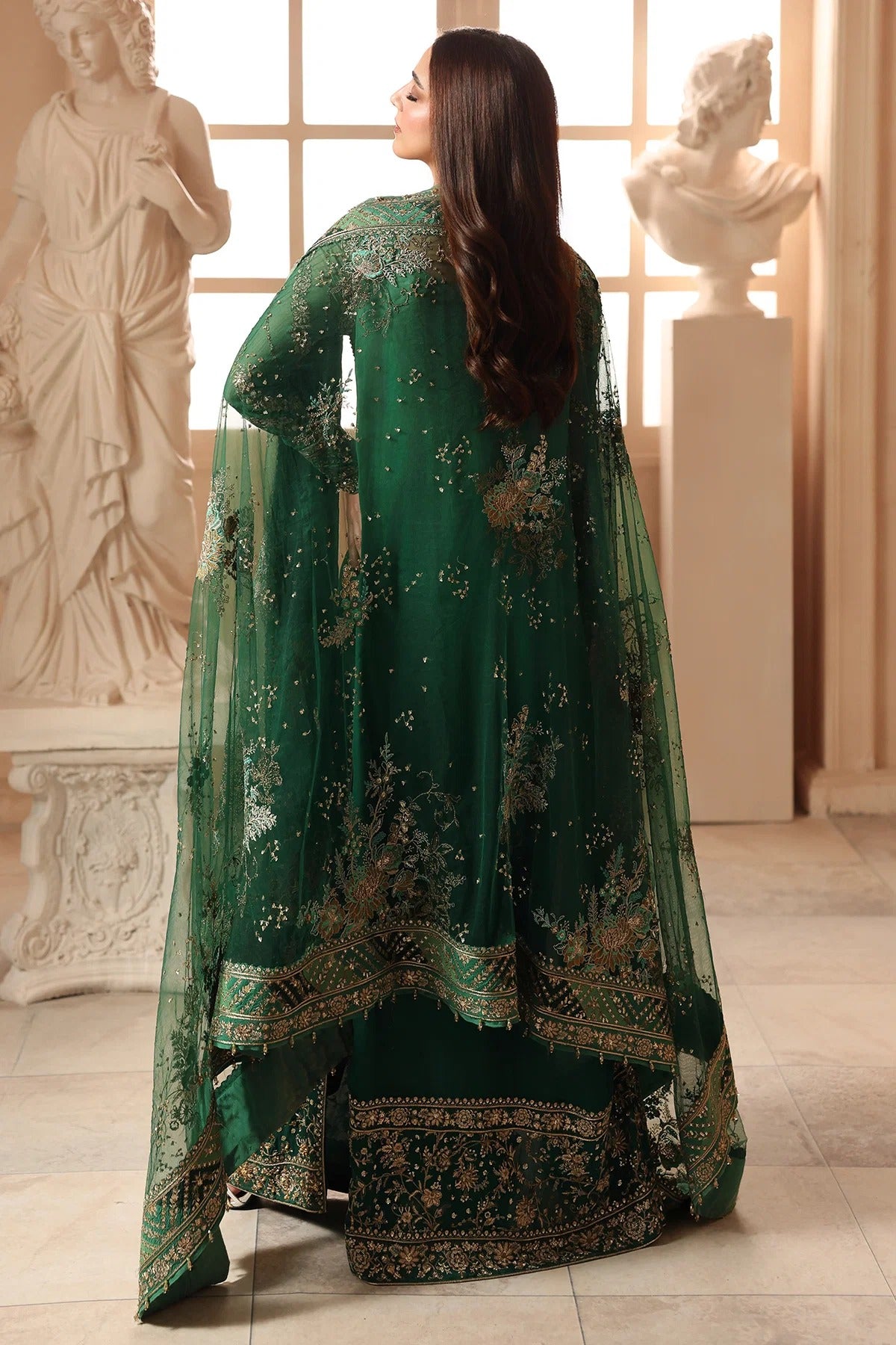 A woman stands with her back to the camera wearing an ornate dark green traditional outfit with intricate gold embroidery and a matching sheer dupatta, photographed in a classical interior with white statue busts.
