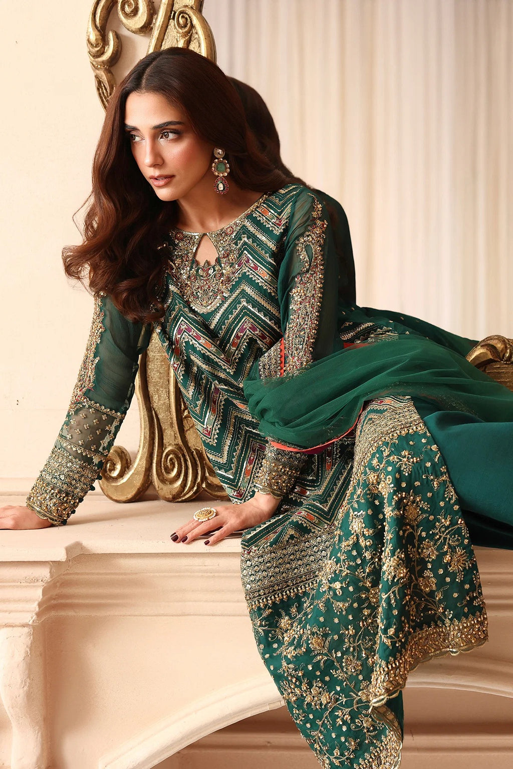 A woman models a dark green traditional South Asian outfit with intricate gold embroidery and geometric patterns, posed on a cream-colored stone ledge in front of a gold-accented chair.