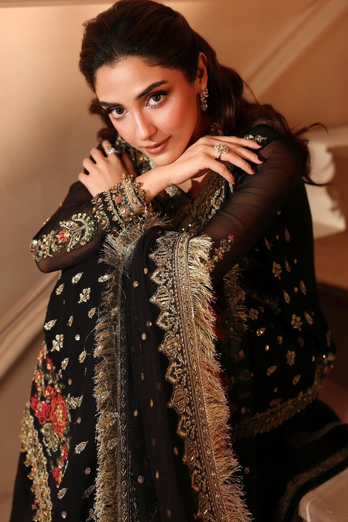 A woman wearing an ornate black traditional outfit with heavy gold embroidery, floral motifs, and a matching sheer dupatta draped over her shoulders, posed indoors against a warm beige wall.