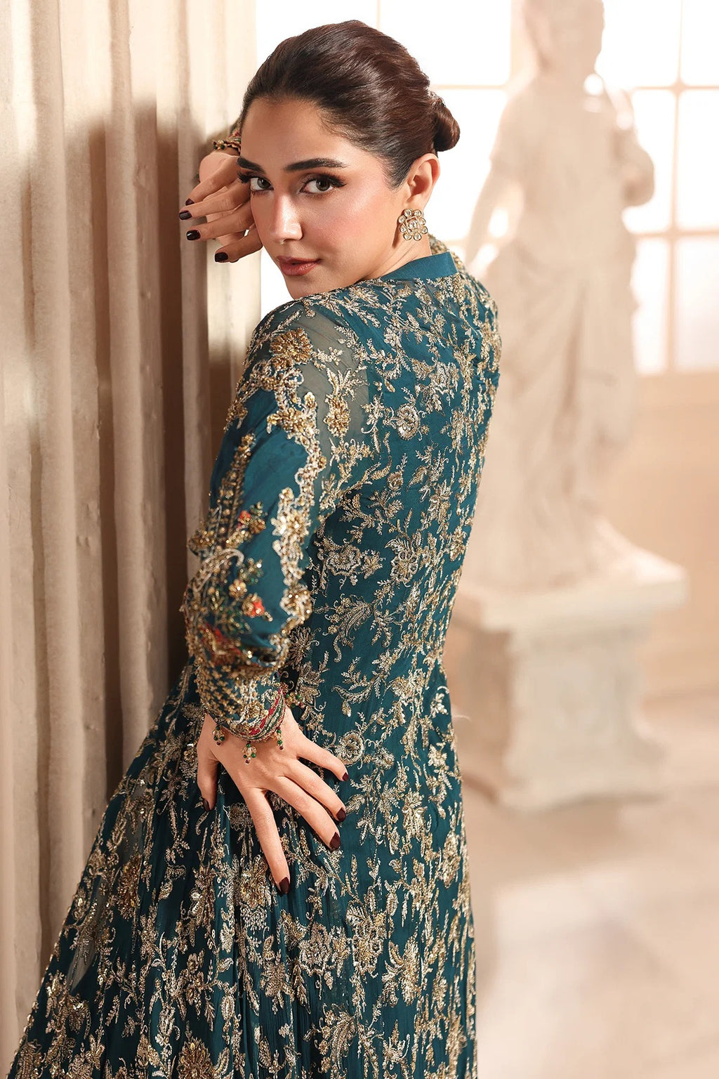 A woman wearing a dark teal embroidered dress with intricate gold and beige floral patterns, posing in an elegant indoor setting with a classical statue in the background.
