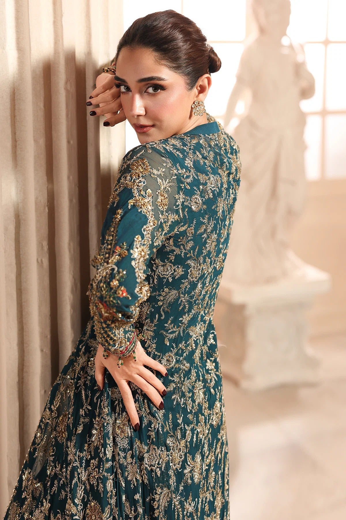 A woman wearing a dark teal embroidered dress with intricate gold and beige floral patterns, posing in an elegant indoor setting with a classical statue in the background.