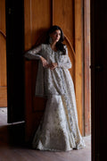 A woman models a traditional South Asian outfit consisting of a silver embroidered kurta and a white mermaid-style skirt with floral embroidery, standing in a doorway with wooden paneling.
