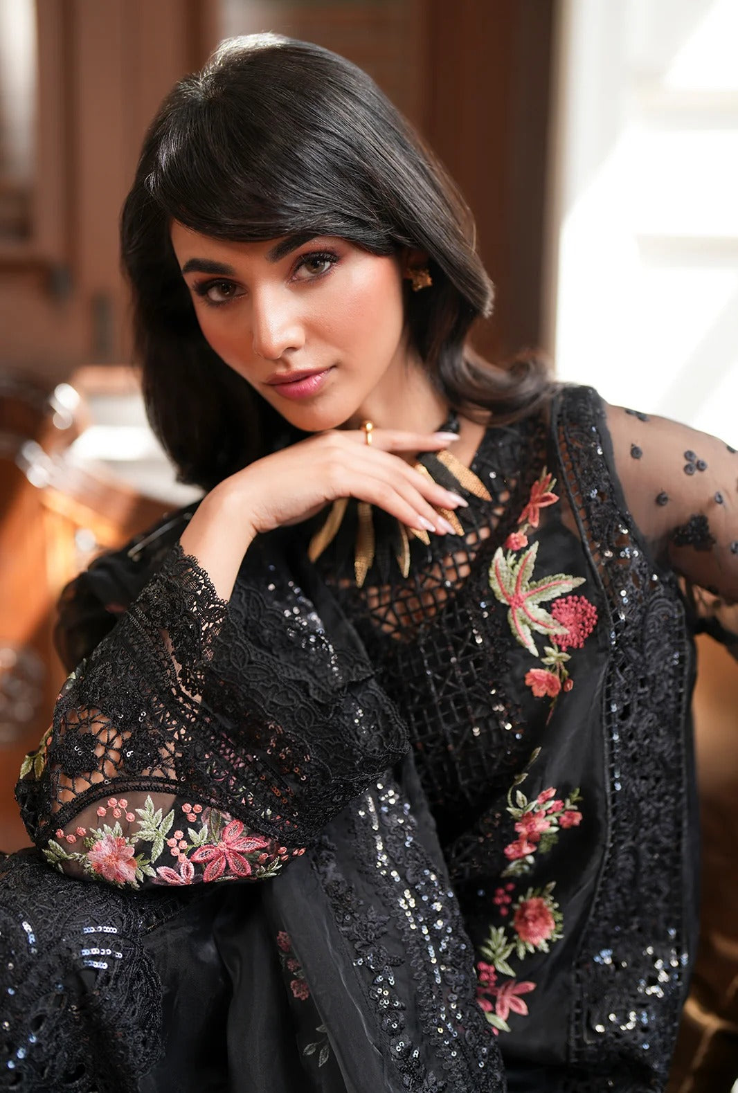 A woman wearing a black embroidered and sequined traditional outfit with sheer sleeves and floral detailing, posed indoors against a softly blurred background.