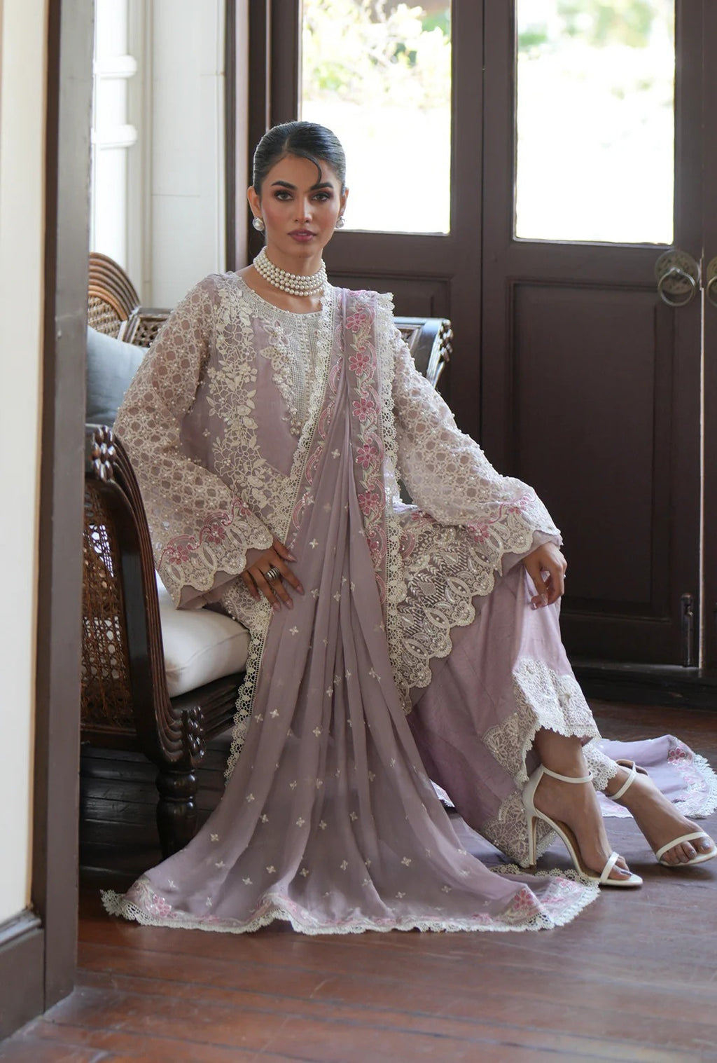 A woman models a traditional Indian salwar kameez outfit in a dusty lavender and beige color palette, featuring intricate floral embroidery, lace borders, and a matching dupatta, set against a background of dark wooden doors and a wicker chair.