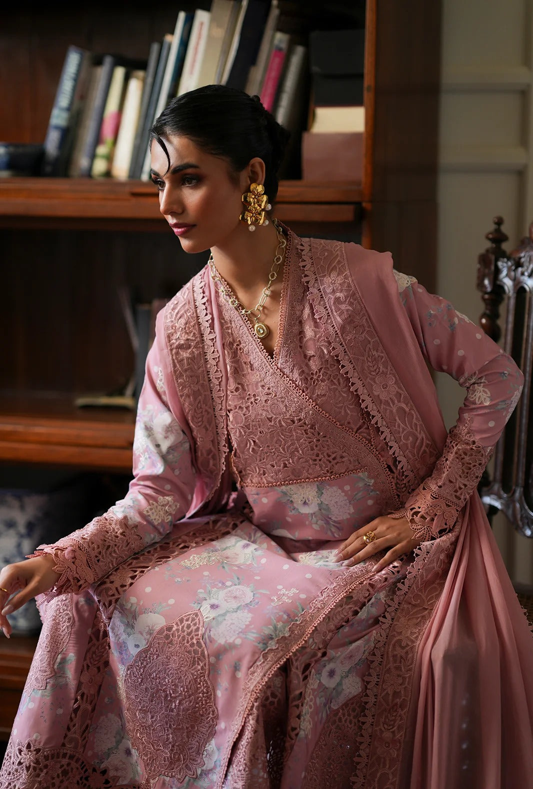 A woman seated indoors wearing a pink embroidered traditional outfit with floral lace detailing, gold jewelry, and a draped matching fabric, set against a bookshelf background.