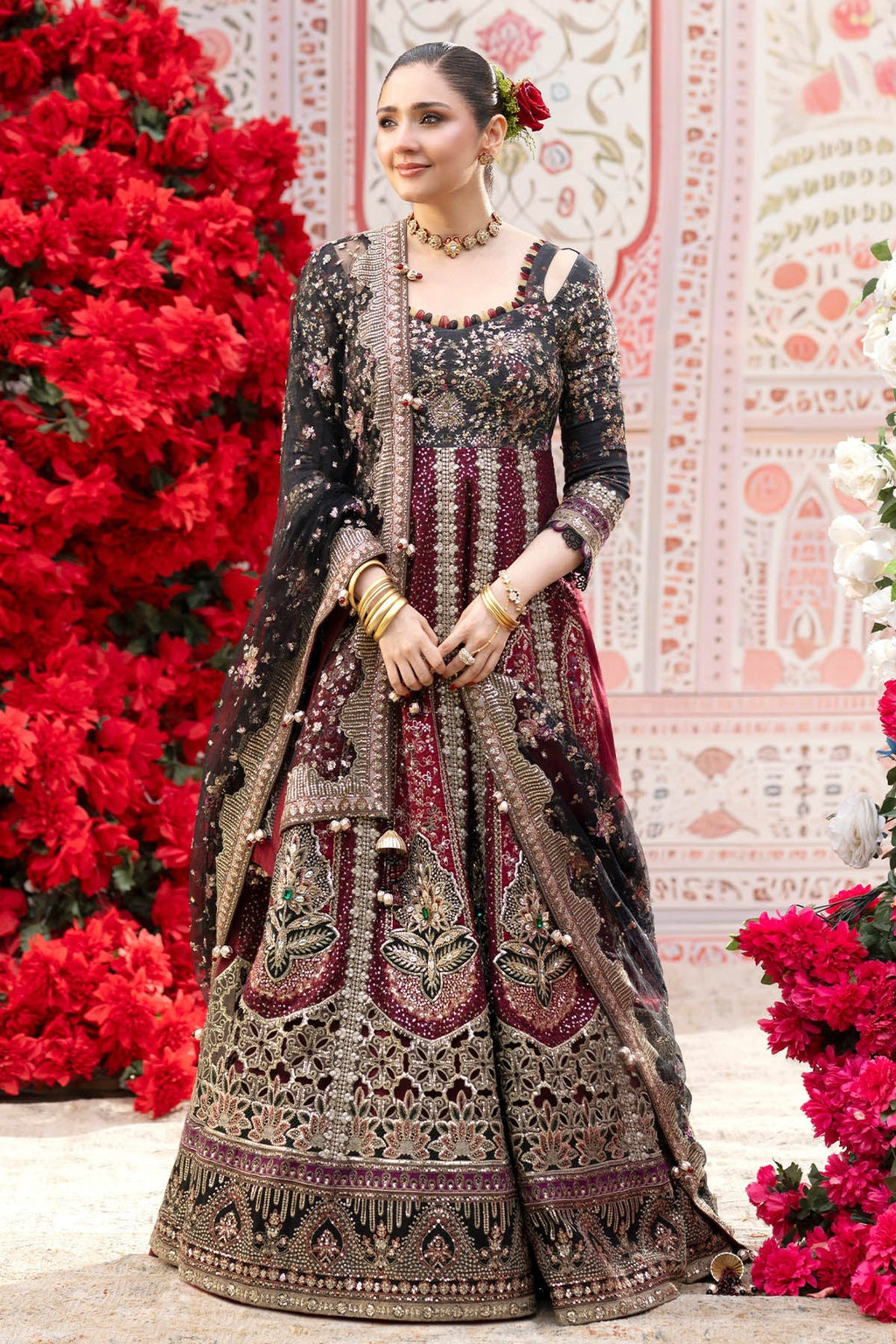 A woman models an ornate black and deep red embroidered traditional dress with a matching dupatta, standing in front of a decorative pink patterned backdrop with red floral arrangements.