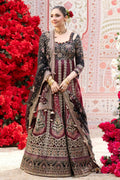 A woman models an ornate black and deep red embroidered traditional dress with a matching dupatta, standing in front of a decorative pink patterned backdrop with red floral arrangements.