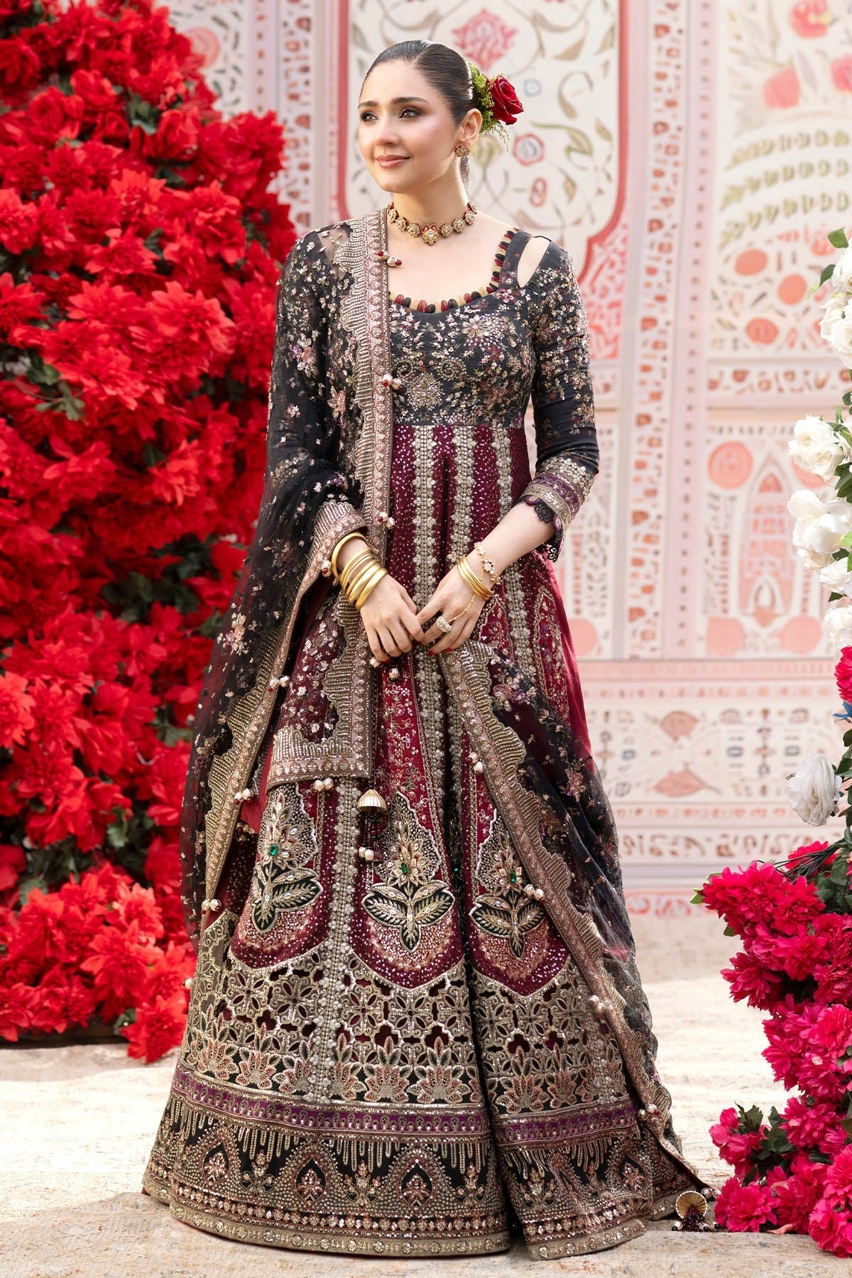 A woman models an ornate black and deep red embroidered traditional dress with a matching dupatta, standing in front of a decorative pink patterned backdrop with red floral arrangements.