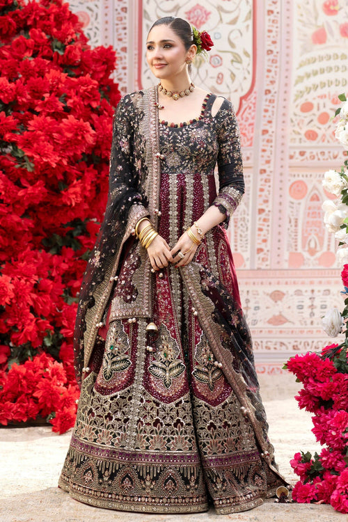 A woman models an ornate black and deep red embroidered traditional dress with a matching dupatta, standing in front of a decorative pink patterned backdrop with red floral arrangements.
