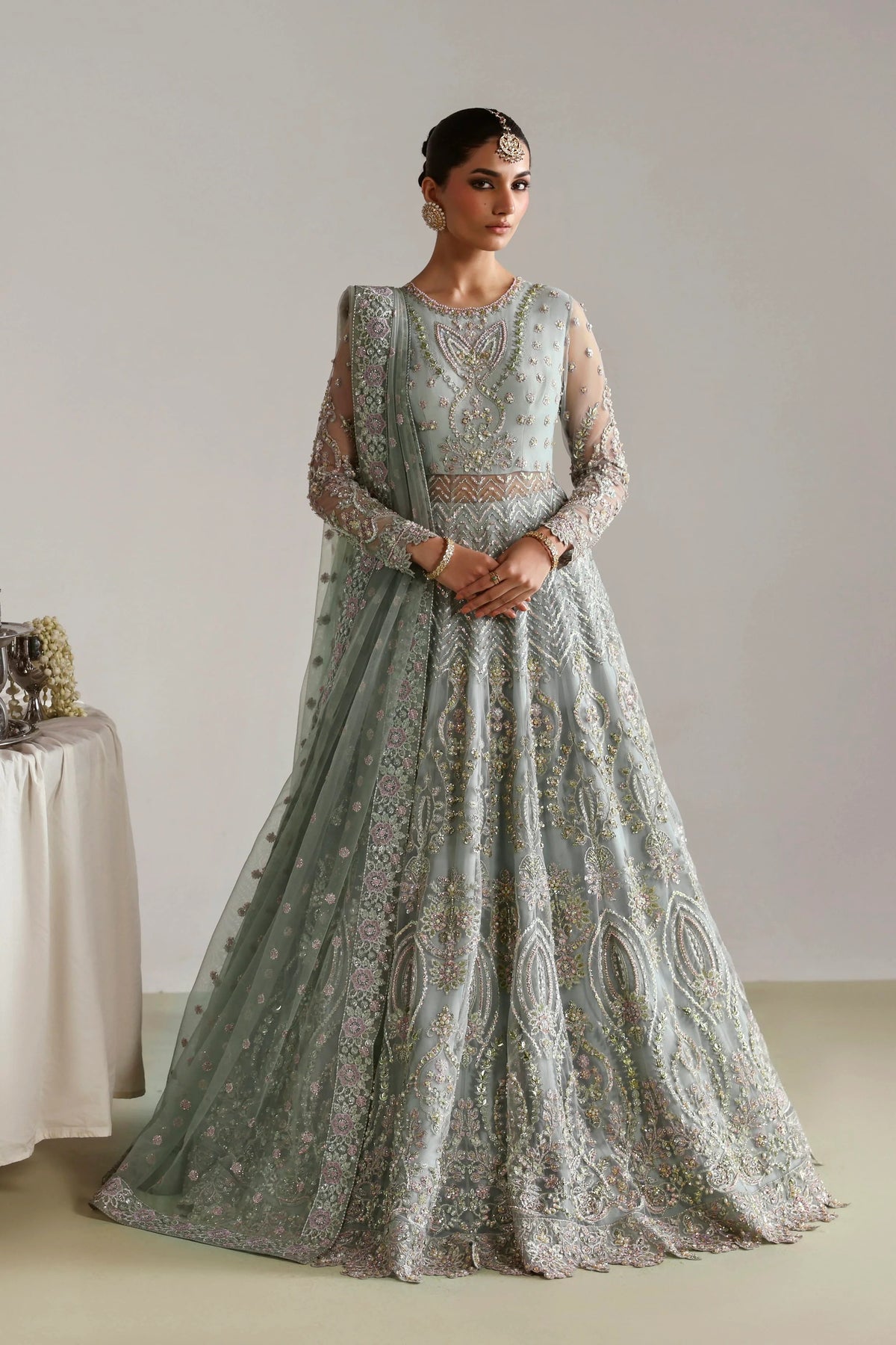 A woman modeling a heavily embroidered light sage green anarkali gown with intricate gold and silver beadwork, set against a neutral studio background.