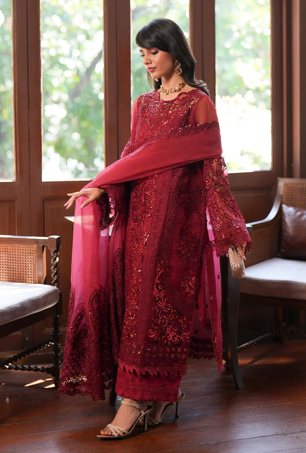 A woman models a deep red embroidered traditional outfit with a matching dupatta in a warm, wood-paneled room. The outfit features intricate floral embroidery and beadwork, and is styled with gold-toned high-heeled sandals.