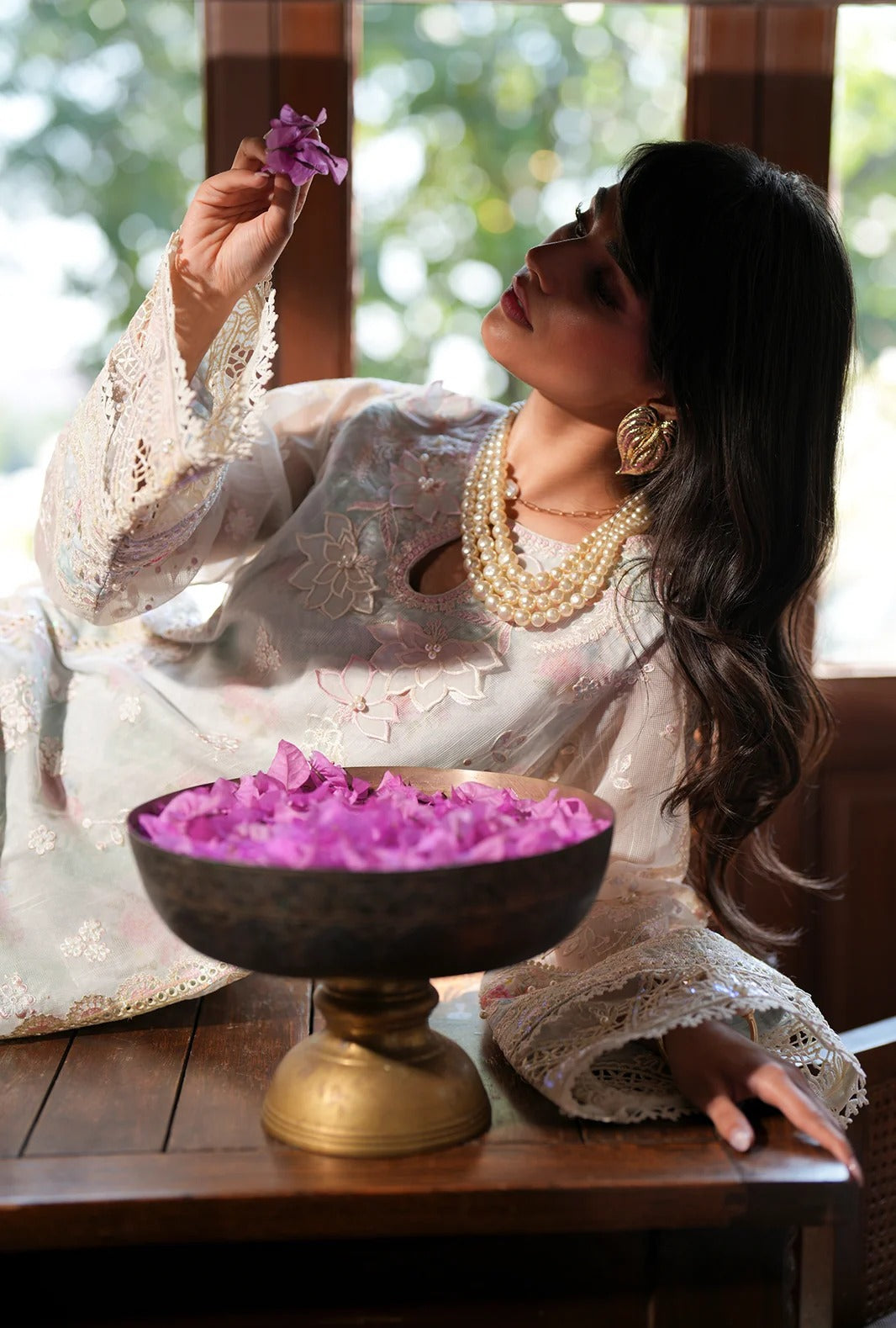 A woman in a white embroidered traditional outfit with lace sleeves holds a purple flower above a bronze pedestal bowl filled with pink petals, set in a rustic indoor environment.