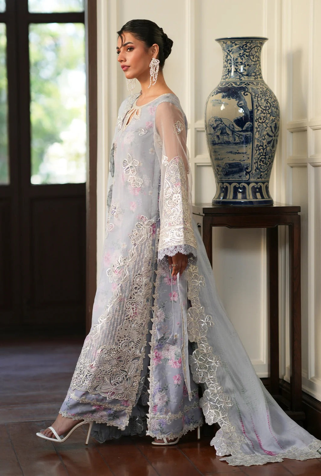 A woman models a light blue-gray embroidered Anarkali gown with matching sheer dupatta in an elegant indoor setting with a blue-and-white ceramic vase on a wooden table.