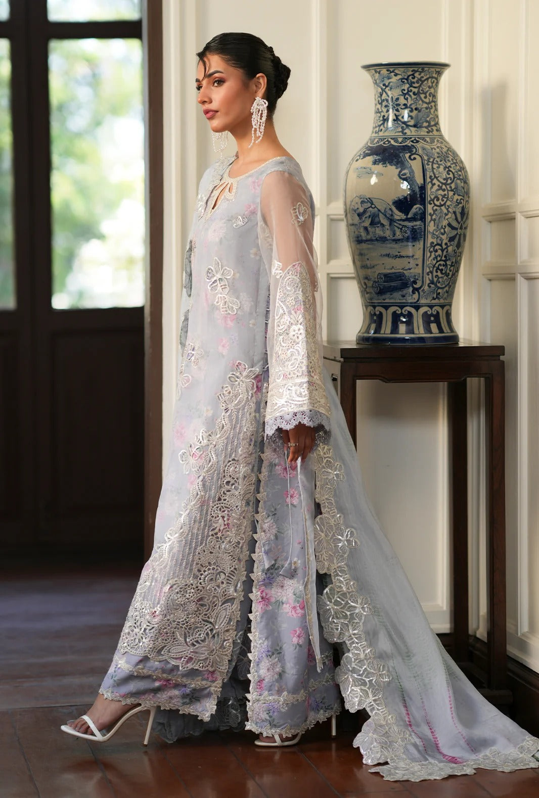 A woman models a light blue-gray embroidered Anarkali gown with matching sheer dupatta in an elegant indoor setting with a blue-and-white ceramic vase on a wooden table.