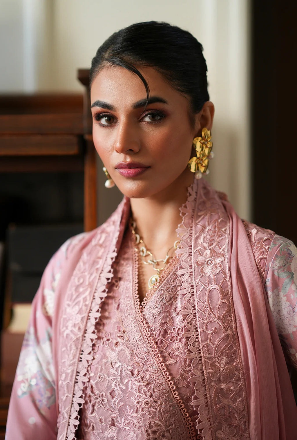 A woman wearing a pink embroidered traditional outfit with matching draped fabric, gold jewelry, and a dark hair styled in a low bun, photographed indoors against a softly blurred background.