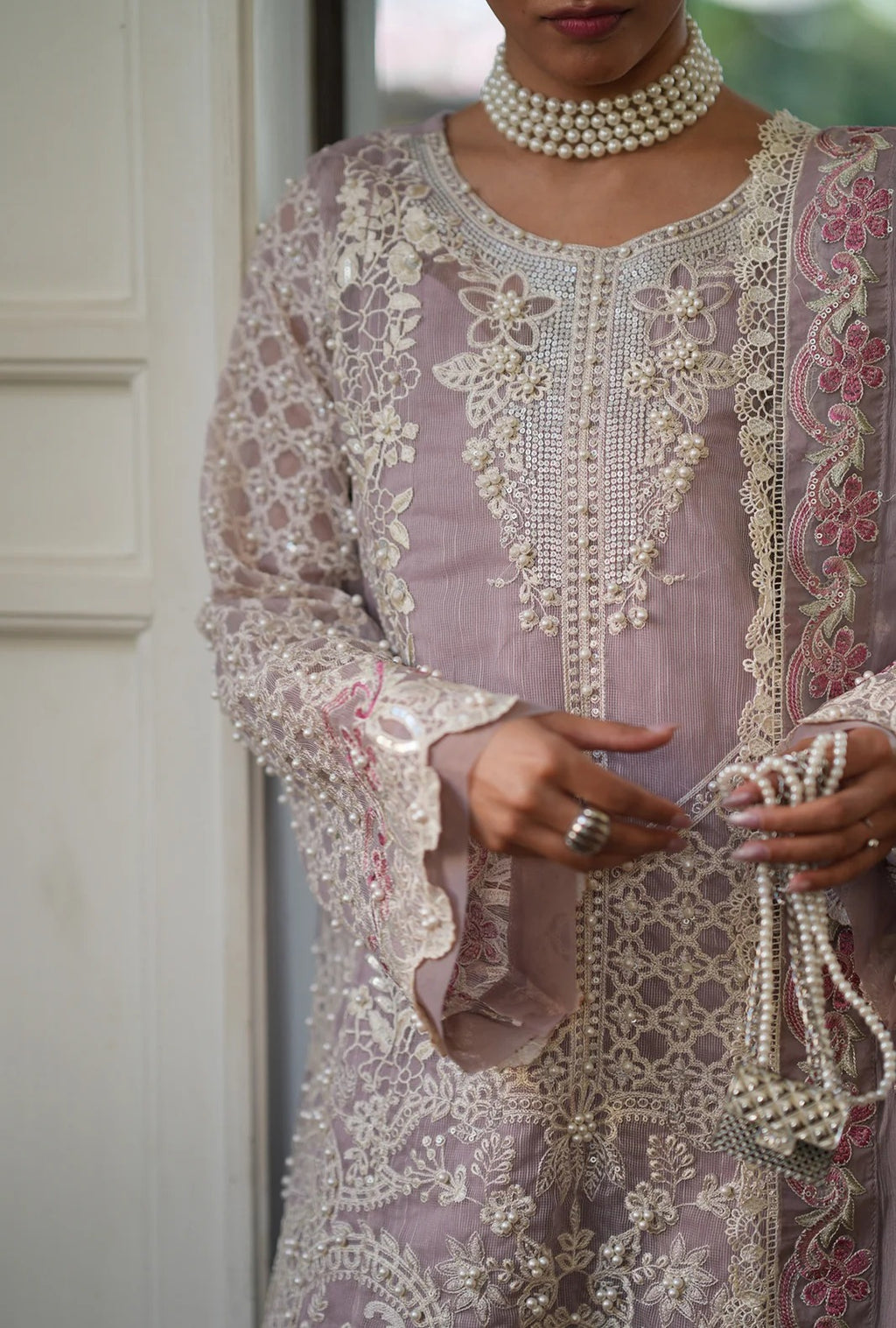A close-up of a person wearing an ornate lavender and beige embroidered traditional outfit with intricate beadwork, floral lace, and pearl necklaces, while holding pearl strands.